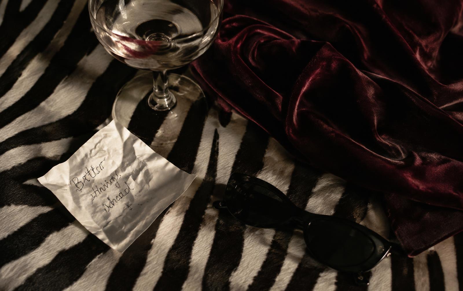 Zerknitterter Zettel, Coupe-Glas mit Lippenstiftabdruck, Samt und Sonnenbrille auf einem Zebrafell